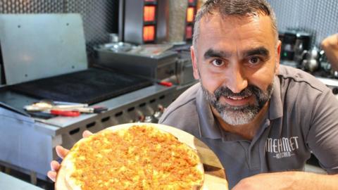 A smiling Mehmet Ulucan shows a plate of food to the camera in his restaurant kitchen. He has grey/black hair and a beard and wears a Mehmet Kitchen polo shirt.