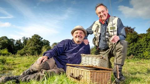 bob lying on grass, with paul knealing and hand on shoulder. picnic basket in foreground