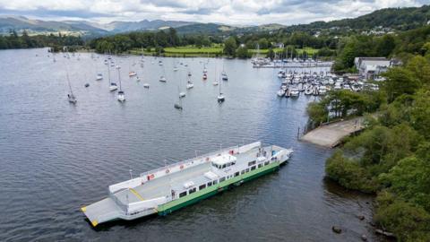 A large grey and green car ferry is on a scenic lake surrounded by other, smaller boats. Windermere is bordered by greenery.