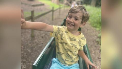 A young boy with blond hair is smiling at the bottom of a green slide and looking to his right with his arm stretched out. He is wearing a yellow top with tigers on it and blue shorts.