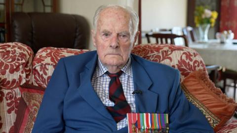 Max looking into the camera and wearing war medals and a tie and jacket.