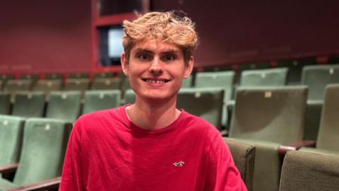 Ciaran O'Brien in a red long-sleeved top, sitting in an audience chair inside Bristol Old Vic