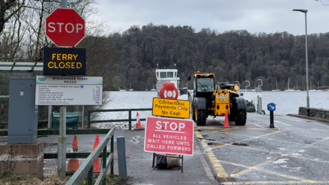 There's grey concrete road in the foreground, and a red sign which reads 'STOP. All vehicles wait here until called forward.' On the left there's a red stop sign, with an illuminated sign underneath saying 'ferry closed.' In the background there is a lake and a forest on the other side.