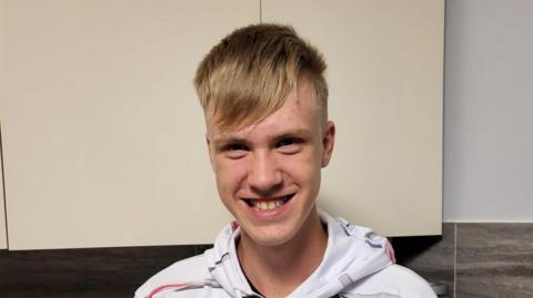 A boy with short blonde hair wearing a white hooded top smiles at the camera