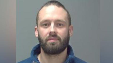 A man with close shaven brown hair and a beard, wearing a blue fleece top.