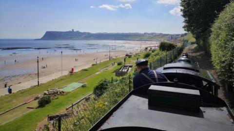 A train driver's head is seen above a miniature train which is travelling along a track above the Scarborough promenade. 