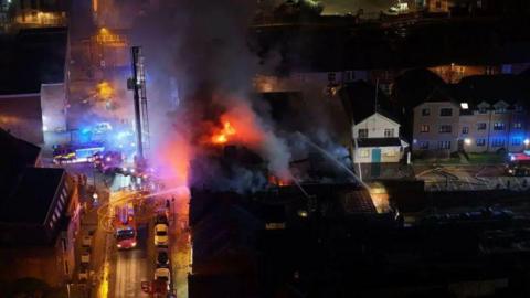 A drone shot of flames and large plumes of smoke coming from a building. There are large amounts of emergency service vehicles in the road next to the building.