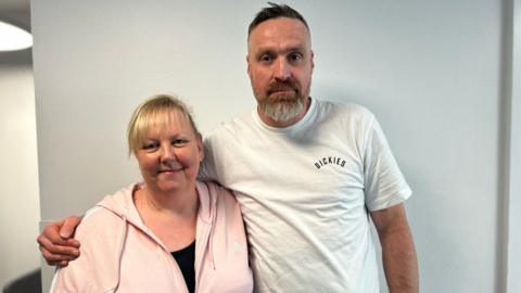 A woman with blonde hair and wearing a pink hoodie, and a man with a grey beard and wearing a white t-shirt, smiling with their arms around each other in front of a plain white wall