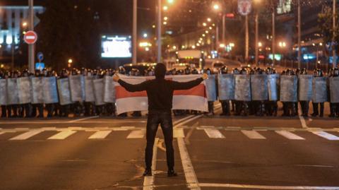 A protest is Belarus, August 2020. A lone man holds a white flag with a red stripe up to a crowd of riot police. 