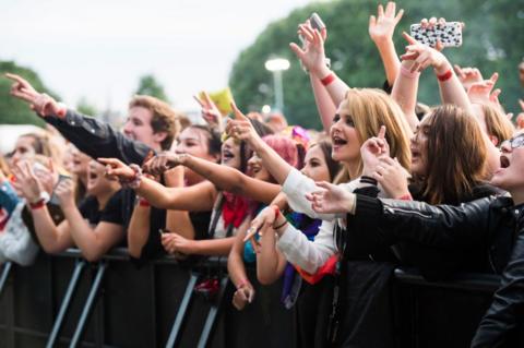 A crowd of people hanging over a barrier at a concert with their hands in the air, singing along. The crowd is mainly made up of young people with different coloured clothing on and different hairstyles.