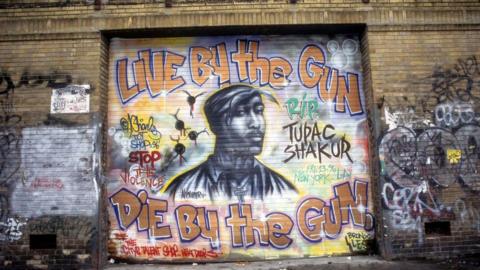 A mural of Tupac shows his face, and the phrase: Live by the gun die by the gun