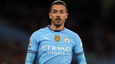 Footballer Jack Grealish runs on a football pitch. He is wearing a sky blue Manchester City jersey with long sleeves underneath it. 