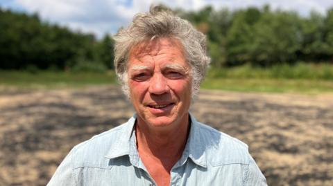 Guy Singh-Watson is wearing a blue short-sleeved shirt as he stands in the bright sunshine in a dry reservoir on a farm at Riverford Organic Farmers in Devon.