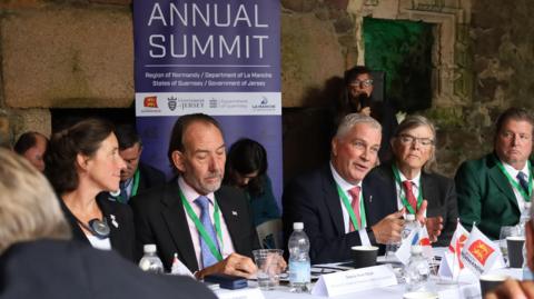 Men and women wearing suits, sit at a table in conversation. Behind them is a purple poster that reads 'annual summit.