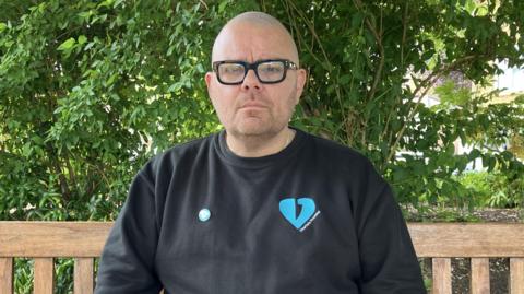 A man, Martin Cosser, sat on a wooden bench wearing a black top. The top is printed with a logo, a blue heart with a knife through it.