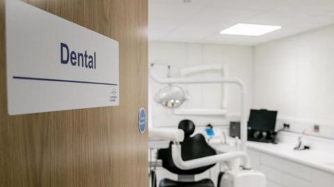 A stock image of a dentist's studio. A door with a signs saying 'dental' opens to a studio with a dentist chair and equipment in the blurred background.