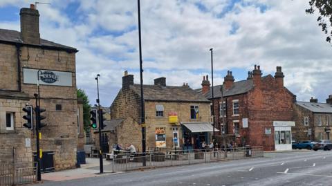 A city street pictured from the other side of a road, featuring bars with punters sat outside. Other street furniture is visible.