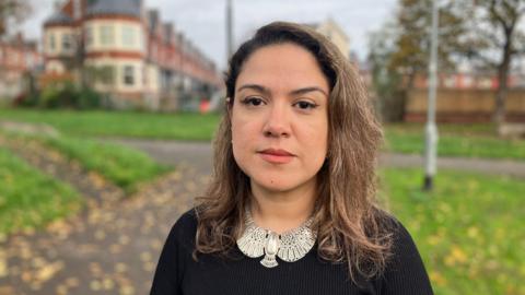 A woman with brown hair stands in a park. She is wearing a black top and large silver jewellery.