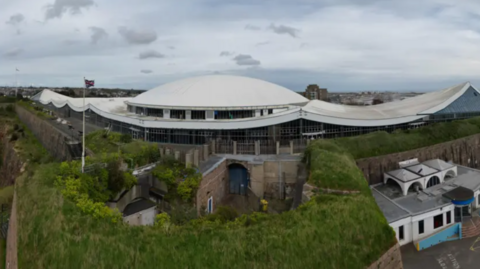 A wide shot of the Fort Regent leisure centre. It is a large white-roofed building surrounded by trees.