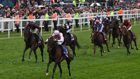 Eight competitors competing at a horse racing event. There is a line of security staff, in orange and yellow hi-vis jackets, standing in front of a crowd. 