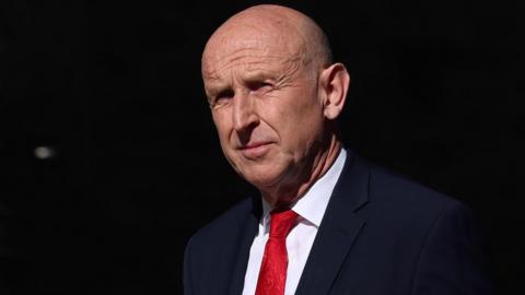 A head and shoulders profile shot of a bald older man in a blue suit, white shirt and deep blue business suit.