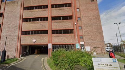 A sign on the right reads "Hook Road", with a blue parking sign above it. There is an exit road from the four-story car park on the left, with a yellow arrow painted on the road and a "no entry" sign on the outside of the building.