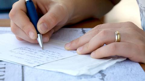 A picture of two hands, one holding a pen and writing on a postcard.