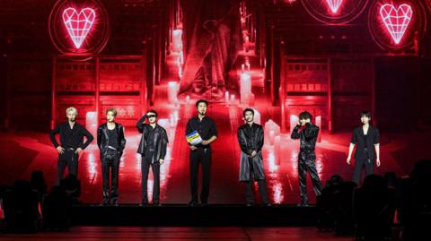 The seven members of K-pop band ENHYPEN on stage at the AO Arena in Manchester. They are all wearing black outfits.