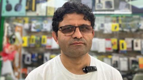 Jabar Farooq looking at the camera, has dark hair and brown eyes. He is wearing a white t-shirt and dark framed glasses. In the background is electronic items in his shop