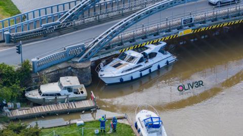 A white cruiser is wedged sideways on underneath a metal bridge.