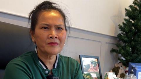A woman frowns as she sits in a shop in front of a white a grey-panelled wall. A small Christmas tree, framed picture and other items can be seen to her right. She is wearing a green polo shirt and silver earrings. 