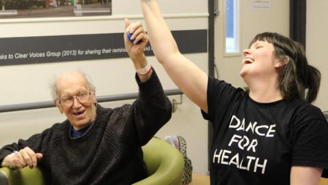A man, sitting in a chair, with one arm raised, smiling. He is wearing glasses and has short grey hair. He has on a jumper. A woman is next to him, laughing, with one arm raised. She is wearing a black T-shirt with "Dance for Health" written on it. Her hair is tied back. 