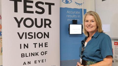 Dr Louise Allen is standing in front of a large sign that says "test your vision in the blink of an eye". There is a tablet on the wall and another sign that says "accurate results in minutes". Dr Allen has shoulder length fair hair and is smiling at the camera. She is wearing a blue blouse and has a name tag around her neck