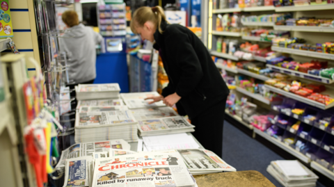 A woman sorts through a pile of newspapers stacked on a table in her newsagents. Behind her are display shelves containing confectionery.