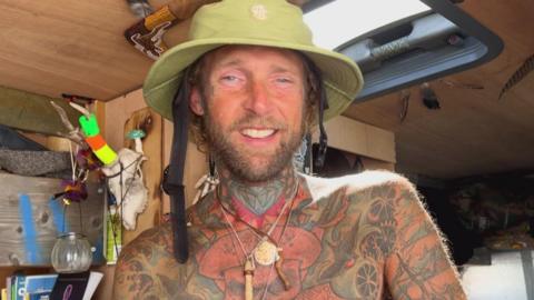 A person sitting inside what appears to be a converted camper van. The person is wearing a light green wide-brimmed hat, several necklaces and has extensive tattoos covering the neck, chest and arms, while the van interior contains wooden shelving, books, hanging ornaments, antlers, a roof skylight and various personal belongings, giving the space a homemade and lived-in feel.