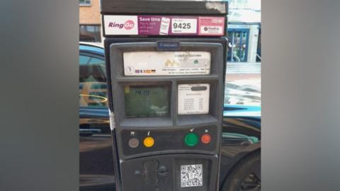 A parking meter in Reading, with a fake QR code on it. It is for on-street parking, with the road behind it.