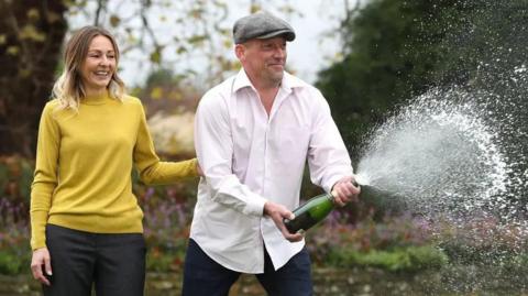 A man in a white shirt and cloth cap opening a bottle of champagne outdoors with spray everywhere next to a woman in a yellow jumper.