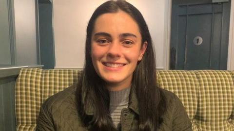 Jenny Hall is white, has long dark hair and smiles at the camera. She has brown eyes and is wearing an olive-coloured jacket.