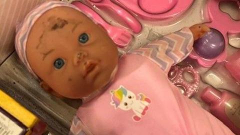 A baby doll in a pink outfit has soot marks on its cheek, nose and forehead. Its surrounded by other pink coloured toys. 