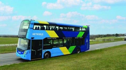 A metrobus hydrogen bus can be seen. It is coloured blue, yellow and green. It is on a quiet road. Either side of there are green fields. 