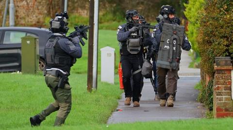 Men in police tactical gear on a footpath surrounded by grass. Their mouths and noses are covered by balaclavas. One is seen carrying a rifle and another carries a shield. Another police officer with a rifle is stood on the grass.