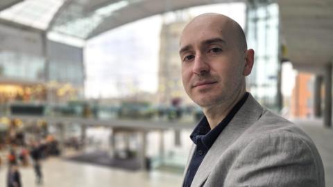 Ben Goldsborough is looking at the camera. He's wearing a grey suit jacket over a dark shirt, and is standing in the Forum in Norwich.