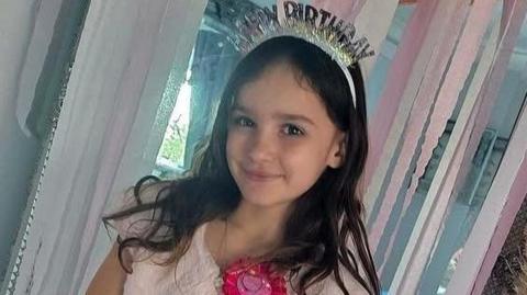 Aria smiles at the camera in front of a doorway filled with pink streamers. She has long brown wavy hair and is wearing a pink dress and shrug. She has a bright pink birthday badge on, and a sparkly headband which reads "HAPPY BIRTHDAY".