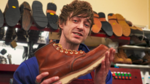 A man with medium length brown hair holding up a brown boot. There are racks of shoes in the background.