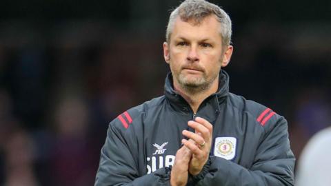 Kenny Lunt applauds the crowd after a game for Crewe. He is wearing a dark grey club coat.