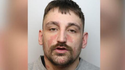 Huckvale stares at the camera with a blank look on his face. He has brown hair, stubble, and a thick moustache and eyebrows. 