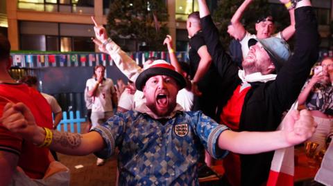 A man wearing a blue England football shirt cheers at the camera with his arms out stretched. He is surrounded by others who are also cheering for joy.