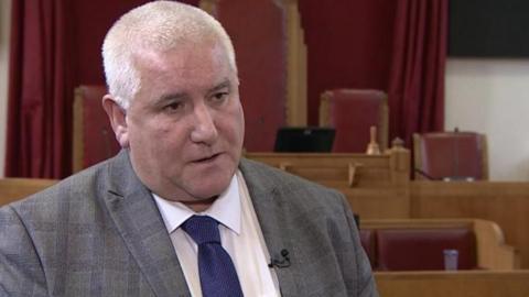 A man stands inside a room, which has wooden chairs with maroon cushions and maroon curtains. He has short grey hair and is wearing a grey suit jacket, a white shirt and a dark blue tie.