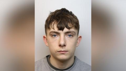 A police custody photo of Ciaran Newman. He has short brown hair and is wearing a grey top.