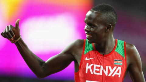 Kipyegon Bett, wearing a red and green athletics vest, gives a 'thumbs up' gesture while looking to his right during a track meeting